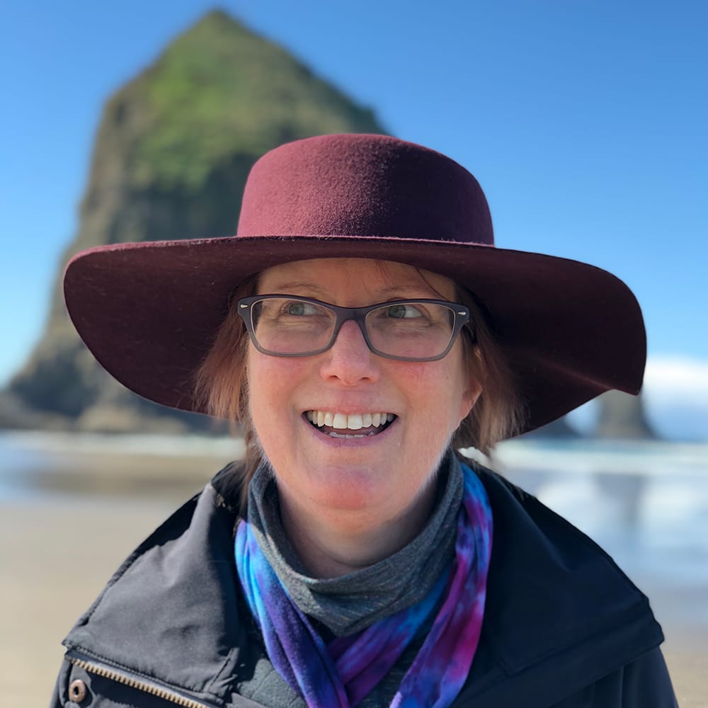 A smiling, beautiful woman wearing glasses and a wide-brimmed hat photographed on vacation at Cannon Beach, Oregon.